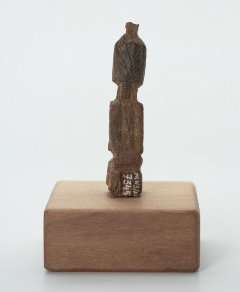 rzeźba - Ujęcie z tyłu; Mała figurka antropomorficzna, najprawdopodobniej przedstawienie przodka. Głowa, rysy twarzy, ręce, nogi wydobyte z bryły drewna płytkimi, ostrymi, prostymi cięciami, co nadaje figurce zgeometryzowana formę. Mogła pełnić rolę osobistego fetysza.