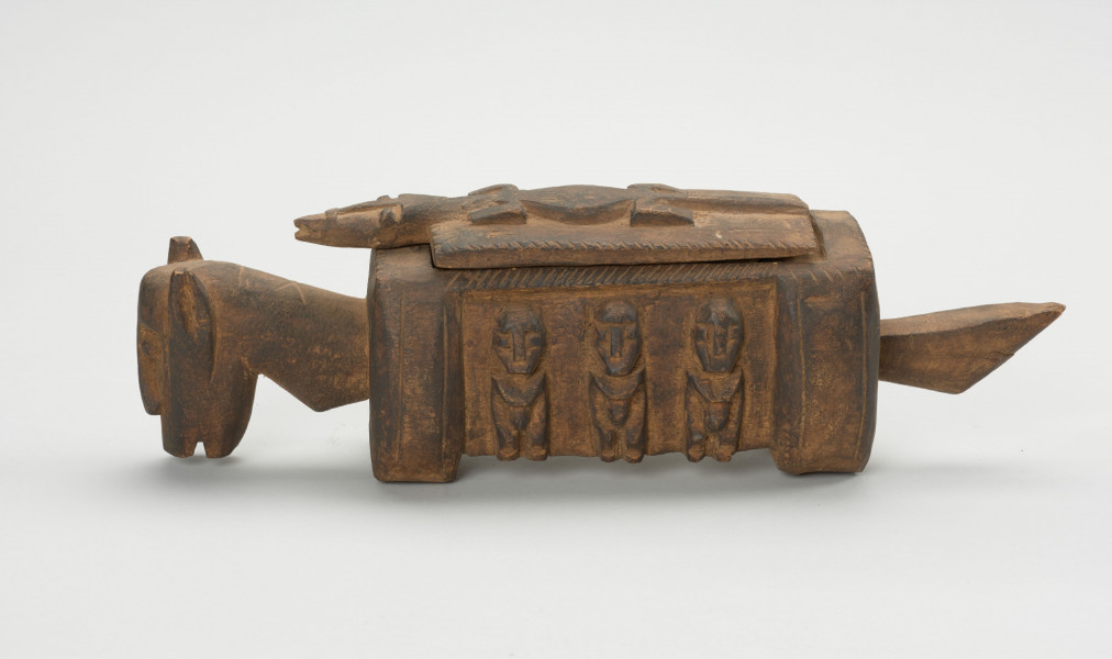 rzeźba - Ujęcie z lewego boku; Arka Nommo zwana inaczej aduno koro lub arką świata, jest wyobrażeniem mitycznej łodzi. Drewniana rzeźa przypominająca konia. Bok zdobiony trzema postaciami w formie płaskorzeźby, stojące w rzędzie. Głowa obiektu zwrócona w stronę ziemi. Na grzbiecie druga część obiektu w formie przykrywki kształtem przypominająca krokodyla na drwnianej prostokatnej podstawie.