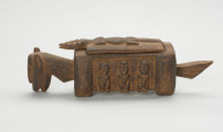 rzeźba - Ujęcie z lewego boku; Arka Nommo zwana inaczej aduno koro lub arką świata, jest wyobrażeniem mitycznej łodzi. Drewniana rzeźa przypominająca konia. Bok zdobiony trzema postaciami w formie płaskorzeźby, stojące w rzędzie. Głowa obiektu zwrócona w stronę ziemi. Na grzbiecie druga część obiektu w formie przykrywki kształtem przypominająca krokodyla na drwnianej prostokatnej podstawie.