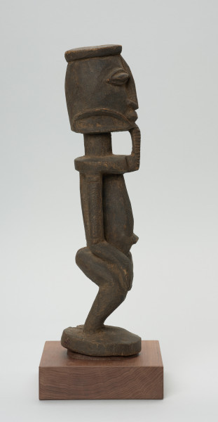 rzeźba - Ujęcie prawego boku. Postać stojąca, z ugiętymi w kolanach nogami. Głowa i tułów wyodrębnione od szyi prosta linią. Oczy, nos, usta wypukłe, broda dekoracyjna sięgająca górnej części tułowia. Ręce przylegające do tułowia, dłonie ułożone na kolanach, brzuch zaokrąglony, z wyraźnie wyodrębnionym pępkiem.