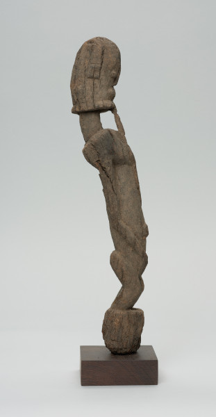 rzeźba - Ujęcie prawego boku. Figura antropomorficzna. Rysy zatarte, widoczna broda, wypukłe, okrągłe oczy. Korpus wydłużony, ręce przylegające do tułowia. Pośladki zaokrąglone. Postać stoi na małym, zaokrąglonym cokoliku.