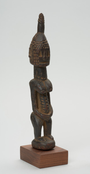 rzeźba - Ujęcie przodu skosem w prawą stronę.  Drewniana figura postaci kobiety. Podłużna głowa ze scheycznie zaznaczonymi rysami i ozdobną wysoką fryzurą. Długa masywna szyja, szczupły tułów. Wyraźnie zaznaczone piersi, długi ręce splecione z przodu. Postać ma krótkie nogi ugięte w kolanach.