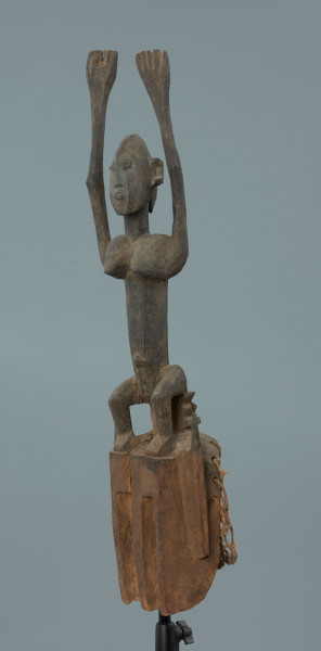 drewniana maska - Ujęcie z przodu, prawej strony; Drewniana, rzeźbiona maska. Nad częścią twarzową, uproszczoną, z wywierconymi dziurami w miejscu oczu, figura postaci ludzkiej z rękoma wzniesionymi do góry. Głowa jajowata, spiczasto zakończona. Zaznaczone uszy. Na wąskim tułowiu wyeksponowane piersi.