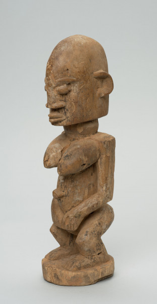 rzeźba - Ujęcie ze skosu z prawej; Figura - postać kobiety w pozycji stojącej. Sylwetka masywna, przysadzista. Głowa duża, z wyrzeźbionymi uszami. Ręce i piersi wydobyte z bryły głębokimi nacięciami. Dłonie ułożone na łonie. Widoczne stopy. Związana z kultem przodków.