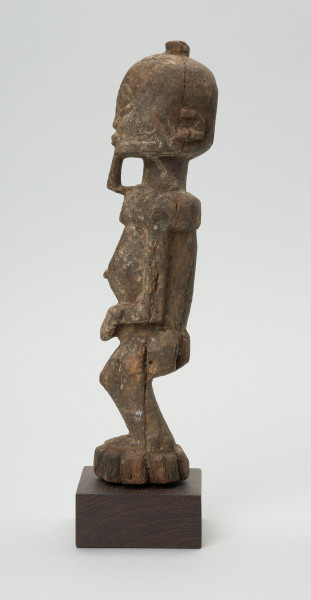 rzeźba - Ujęcie lewego boku. Figura przedstawiająca mężczyznę stojącego na nogach ugiętych w kolanach. Głowa duża, zaokrąglona, oczy duże, ze źrenicami, wyraźnie zaznaczona bródka.