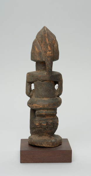 rzeźba - Ujęcie z tyłu. Figura przedstawiająca postać kobiety siedzącej na stołku. Postać masywna, głowa nieproporcjonalnie duża, ramiona proste, ręce przylegające do tułowia, dłonie spoczywają na kolanach. Nos długi, prosty, w kształcie strzałki, oczy i usta zaznaczone symbolicznie.  W dolnej części widnieje numer inwentarzowy.