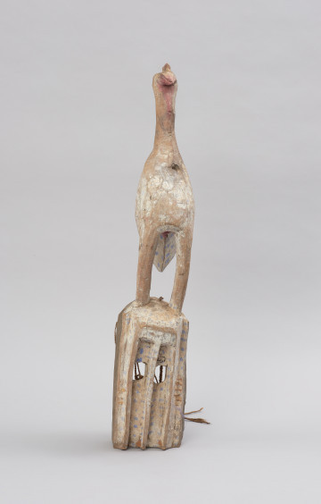 Drewniana maska z figurą ptaka - Ujęcie z przodu. Drewniana maska z figurą ptaka. Część twarzowa maski prostokątna, a wyciętymi kwadratowymi oczami. Na szczycie części twarzowej przedstawienie ptaka stojącego na długich nogach. Całość polichromowana.