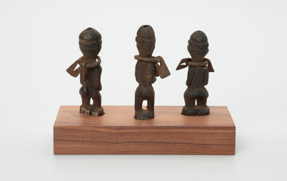 rzeźba - Ujęcie z tyłu. Zestaw składa się z trzech małych, drewnianych figurek w pozycji stojącej przedstawiających postacie ludzkie wyobrażające przodków. Rzeźbom nie zaznaczono rysów twarzy, na szyi ozdobiono je żelaznym pierścieniem z podwójnym żelaznym wisiorkiem o trapezowatym kształcie.