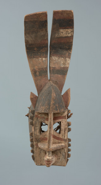 Drewniana maska z długimi płaskimi rogami - Ujęcie z przodu; Drewniana maska. Część twarzowa w formie zaokrąglonego trójkąta równoramiennego. Oczy wycięte, trapezowe. Nos prosty, odstający, nozdrza cienkie, wypełniające całą szerokość części twarzowej. Na górze małe uszy oraz duże płaskie rogi.