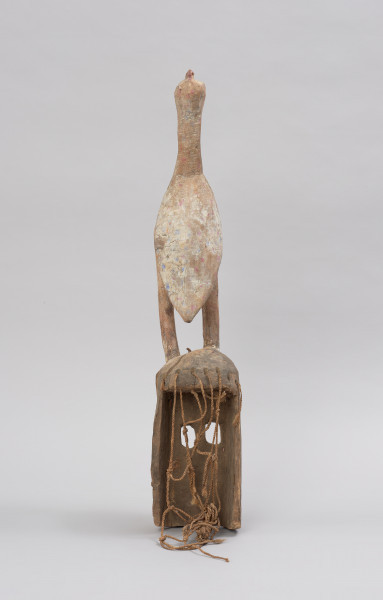 Drewniana maska z figurą ptaka - Ujęcie z tyłu. Drewniana maska z figurą ptaka. Część twarzowa maski prostokątna, a wyciętymi kwadratowymi oczami. Na szczycie części twarzowej przedstawienie ptaka stojącego na długich nogach. Całość polichromowana.