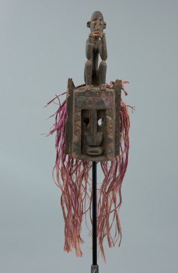 drewniana, rzeźbiona maska - Ujęcie z przodu. Drewniana, rzeźbiona maska małpy. Część twarzowa prostokątna, oczy wycięte, trójkątne. Nos i usta zaznaczone schematycznie. Na szczycie postać małpy w pozycji siedzącej, z nogami podkulonymi, rękami opartymi na kolanach. W dłoniach i ustach trzyma patyk.