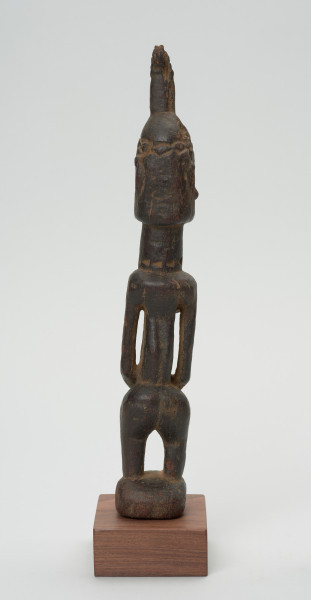 rzeźba - Ujęcie z tyłu. Drewniana figura postaci kobiety. Podłużna głowa ze scheycznie zaznaczonymi rysami i ozdobną wysoką fryzurą. Długa masywna szyja, szczupły tułów. Wyraźnie zaznaczone piersi, długi ręce splecione z przodu. Postać ma krótkie nogi ugięte w kolanach.