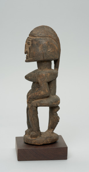 rzeźba - Ujęcie z prawej strony. Figura przedstawiająca postać kobiety siedzącej na stołku. Postać masywna, głowa nieproporcjonalnie duża, ramiona proste, ręce przylegające do tułowia, dłonie spoczywają na kolanach. Nos długi, prosty, w kształcie strzałki, oczy i usta zaznaczone symbolicznie.