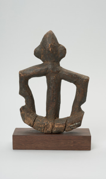 rzeźba - Ujęcie tyłu. Figurka przedstawiająca postać ludzką. Ręce i nogi w przysiadzie wąskie, połączone ze sobą w formę kwadratu. Głowa spiczasta z odstającymi szerokimi uszami. Tułów cienki, prosty.