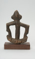 rzeźba - Ujęcie tyłu. Figurka przedstawiająca postać ludzką. Ręce i nogi w przysiadzie wąskie, połączone ze sobą w formę kwadratu. Głowa spiczasta z odstającymi szerokimi uszami. Tułów cienki, prosty.