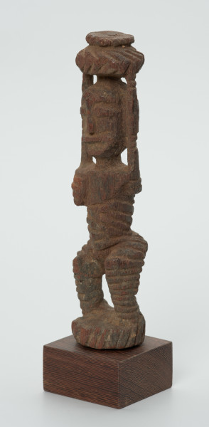 drewniana figura kobiety - Ujęcie z przodu skosem w lewą stronę. Drewniana, rzeźbiona figura kobiety niosącej na głowie naczynie. Postać schematyczna, z uniesionymi ramionami. Głowa jajowata, rysy twarzy uproszczone. Stopy złączone w okrągły cokolik.