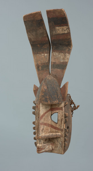Drewniana maska z długimi płaskimi rogami - Ujęcie ze skosu z lewej; Drewniana maska. Część twarzowa w formie zaokrąglonego trójkąta równoramiennego. Oczy wycięte, trapezowe. Nos prosty, odstający, nozdrza cienkie, wypełniające całą szerokość części twarzowej. Na górze małe uszy oraz duże płaskie rogi.
