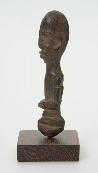 Drewniana figurka z dużą głową i wąskimi oczami - Ujęcie z lewej strony; mała drewniana figurka antropomorficzna. Głowa duża zaokrąglona. Wąskie oczy. Rysy, ręce wydobyte z bryły drewna płytkimi cięciami. Brak nóg.