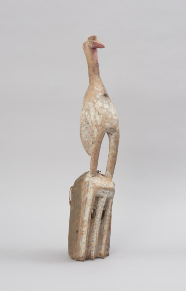 Drewniana maska z figurą ptaka - Ujęcie z przodu z prawej strony. Drewniana maska z figurą ptaka. Część twarzowa maski prostokątna, a wyciętymi kwadratowymi oczami. Na szczycie części twarzowej przedstawienie ptaka stojącego na długich nogach. Całość polichromowana.