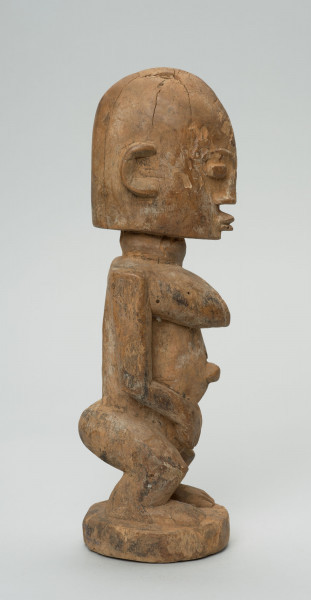 rzeźba - Ujęcie z boku; Figura - postać kobiety w pozycji stojącej. Sylwetka masywna, przysadzista. Głowa duża, z wyrzeźbionymi uszami. Ręce i piersi wydobyte z bryły głębokimi nacięciami. Dłonie ułożone na łonie. Widoczne stopy. Związana z kultem przodków.