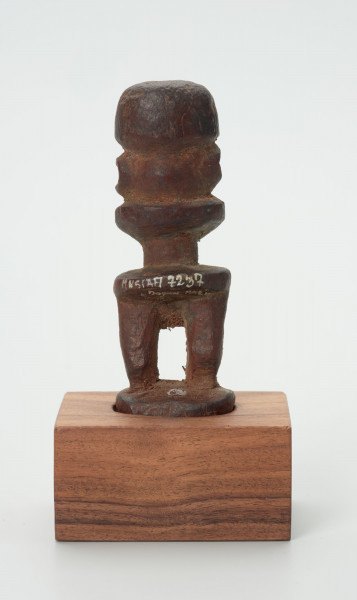 Drewniana figurka postaci ludzkiej z dużą głową i krótkimi nogami. - Ujęcie z tyłu; drewniana figurka postaci ludzkiej. Głowa zajmuje większość powierzchni rzeźby. Brak tułowia. Krótkie nogi. Figura usytuowana na drewnianej podstawie.