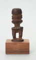 Drewniana figurka postaci ludzkiej z dużą głową i krótkimi nogami. - Ujęcie z tyłu; drewniana figurka postaci ludzkiej. Głowa zajmuje większość powierzchni rzeźby. Brak tułowia. Krótkie nogi. Figura usytuowana na drewnianej podstawie.
