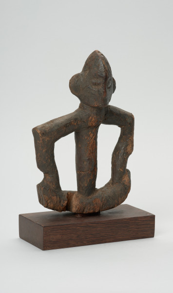 rzeźba - Ujęcie przodu skosem w prawą stronę. Figurka przedstawiająca postać ludzką. Ręce i nogi w przysiadzie wąskie, połączone ze sobą w formę kwadratu. Głowa spiczasta z odstającymi szerokimi uszami. Tułów cienki, prosty.