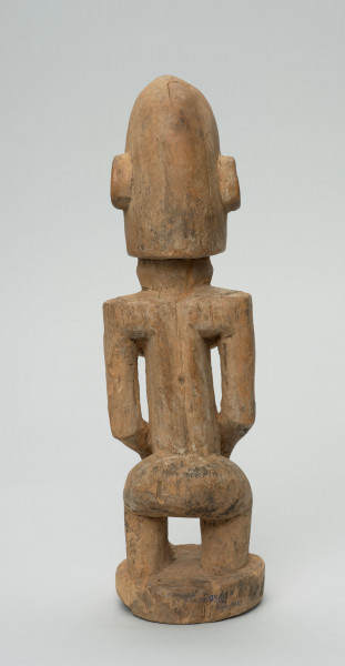 rzeźba - Ujęcie z tyłu; Figura - postać kobiety w pozycji stojącej. Sylwetka masywna, przysadzista. Głowa duża, z wyrzeźbionymi uszami. Ręce i piersi wydobyte z bryły głębokimi nacięciami. Dłonie ułożone na łonie. Widoczne stopy. Związana z kultem przodków.