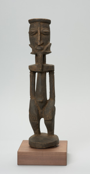 rzeźba - Ujęcie z przodu. Postać stojąca, z ugiętymi w kolanach nogami. Głowa i tułów wyodrębnione od szyi prosta linią. Oczy, nos, usta wypukłe, broda dekoracyjna sięgająca górnej części tułowia. Ręce przylegające do tułowia, dłonie ułożone na kolanach, brzuch zaokrąglony, z wyraźnie wyodrębnionym pępkiem.