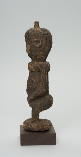 rzeźba - Ujęcie z prawej strony. Figurka - postać kobiety w pozycji stojącej. Głowa duża wychodząca bezpośrednio z tułowia. Ręce przylegające do tułowia. Na głowie mała wypustka.