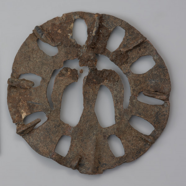 plakietka ażurowa z ptakiem - Ujęcie tyłu plakietki z góry. Metalowa, okrągła plakietka ażurowa z wyobrażeniem ptaka w części centralnej, otoczonego koncentrycznymi kręgami z owalnymi otworami nadającymi całości pewnej lekkości. Na odwrocie plastyczne zeberka ułożone promieniście, wzmacniające konstrukcję.