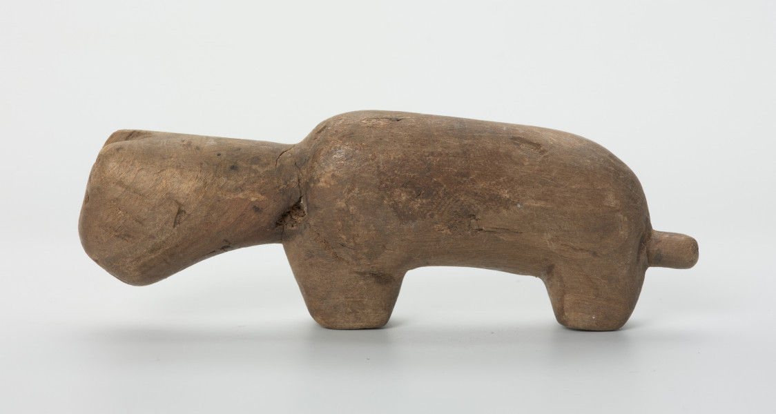 Drewniana figura zwierzęcia z krótkim ogonem - Ujęcie z lewej strony. Drewniana figurka nieokreślonego zwierzęcia z długą szyją, podłużną głową i krótkim ogonem.