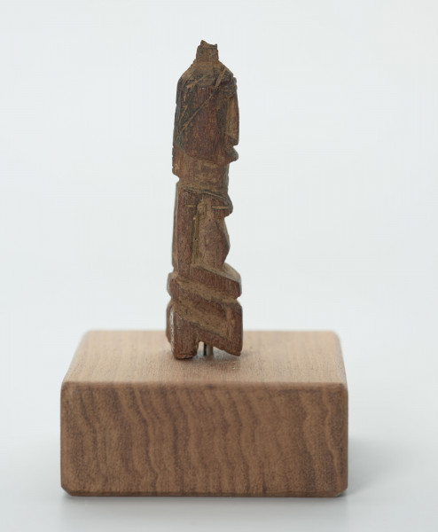 rzeźba - Ujęcie z lewego boku; Mała figurka antropomorficzna, najprawdopodobniej przedstawienie przodka. Głowa, rysy twarzy, ręce, nogi wydobyte z bryły drewna płytkimi, ostrymi, prostymi cięciami, co nadaje figurce zgeometryzowana formę. Mogła pełnić rolę osobistego fetysza.