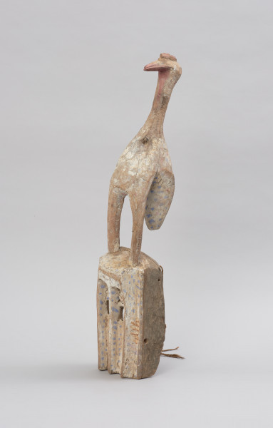 Drewniana maska z figurą ptaka - Ujęcie z przodu z lewej strony. Drewniana maska z figurą ptaka. Część twarzowa maski prostokątna, a wyciętymi kwadratowymi oczami. Na szczycie części twarzowej przedstawienie ptaka stojącego na długich nogach. Całość polichromowana.