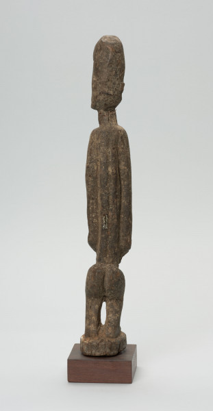 rzeźba - Ujęcie z tyłu; Figura - postać kobiety w pozycji stojącej. Głowa oddzielona od szyi prostą linią. Okrągłe czoło, nos długi, prosty, strzałkowaty, usta i oczy szerokie. Ręce wydobyte płytkimi nacięciami z bryły korpusu. Dłonie spoczywają na łonie. Nogi ugięte w kolanach. Zaznaczone stopy.
