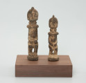 rzeźba - Ujęcie z przodu; Zestaw dwóch małych, drewnianych figurek postaci ludzkich w pozycji stojącej, pokrytych warstwą białej i miedzianej substancji. Figurki mają fryzury lub nakrycia głowy postawione na czubku głowy. Widoczne niewyraźne, okrągłe oczy i linia nosa. Ręce rzeźb  przylegają do tułowia, ich dłonie oparto o lekko wypukły i wystający brzuch, nogi krótkie i ugięte w kolanach, stopy połączone, stoją na okrągłych podstawkach. Obie figurki zostały umieszczone przez konserwatora na wspólnej prostokątnej, drewnianej, ekspozycyjnej podstawie.