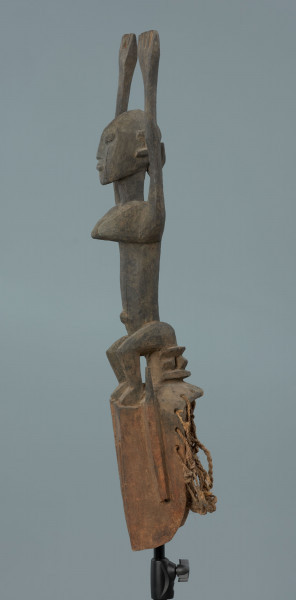 drewniana maska - Ujęcie z prawego boku; Drewniana, rzeźbiona maska. Nad częścią twarzową, uproszczoną, z wywierconymi dziurami w miejscu oczu, figura postaci ludzkiej z rękoma wzniesionymi do góry. Głowa jajowata, spiczasto zakończona. Zaznaczone uszy. Na wąskim tułowiu wyeksponowane piersi.