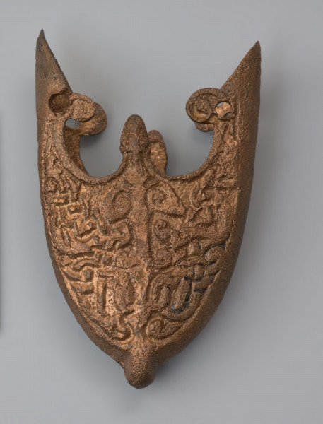 trzewik pochwy miecza typu VI (tzw. grupa gotlandzka) - Ujęcie z przodu. Dobrze zachowane dolne okucie pochwy miecza, z niewielkim ubytkiem po jednej stronie i ogólnym wytarciem powierzchni. Na obu bokach widoczne są szwy odlewnicze. Górna część trzewika zaopatrzona jest w trzy występy, dwa boczne ozdobione wypustkami są wyższe od środkowego. Występ środkowy zwieńczony jest stylizowaną główką zwierzęcą. Dolna strona zaopatrzona jest w centralnie umieszczony występ. Obie strony powierzchni trzewika wypełnia skomplikowany ornament, słabo czytelny w wyniku częściowego wytarcia. Przedstawia on drzewo życia z parą stylizowanych ptaków o długich ogonach oraz u podstawy pnia wijące się ósemkowato mocno wydłużone ciała wężów-smoków.