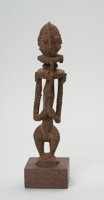 rzeźba - Ujęcie z przodu. Postać o cechach antropomorficznych w pozycji stojącej. Głowa duża, oczy, nos wypukłe. Na wysokości szyi rodzaj kryzy. Ręce pełnoplastyczne, dłonie połączone, spoczywają nad pępkiem. Nogi krótkie. Część górna ozdobiona drobnymi nacięciami.