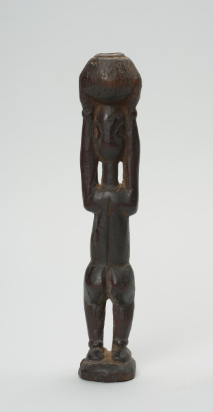 rzeźba - Ujęcie z tyłu. Figura - postać kobiety w pozycji stojącej z rękoma wyciągniętymi ku górze, którymi podtrzymuje stojące na głowie naczynie. Na brzuchu delikatne nacięcia - skaryfikacja. Zaznaczone stopy. Przy dłoniach i stopach - okrągłe zgrubienia - wizerunki bransolet.