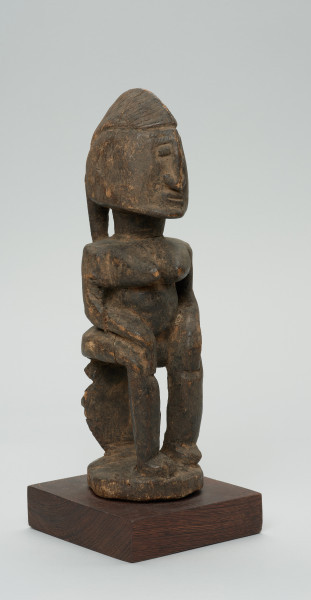 rzeźba - Ujęcie przodu skosem w prawą stronę. Figura przedstawiająca postać kobiety siedzącej na stołku. Postać masywna, głowa nieproporcjonalnie duża, ramiona proste, ręce przylegające do tułowia, dłonie spoczywają na kolanach. Nos długi, prosty, w kształcie strzałki, oczy i usta zaznaczone symbolicznie.