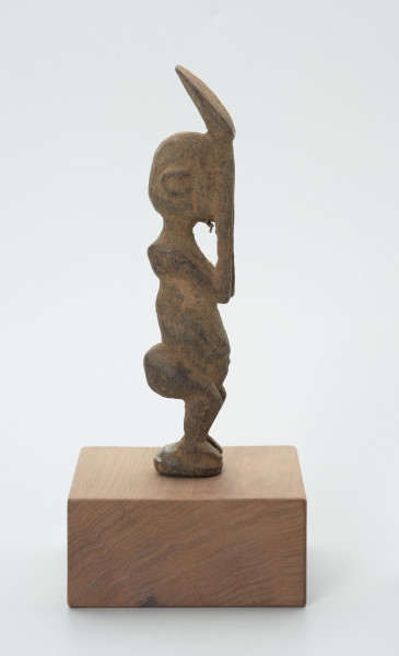 rzeźba - Ujęcie lewej strony. Figura - postać mężczyzny w pozycji stojącej z ugiętym kolanami. Ręce i dłonie nieproporcjonalnie długie/duże w stosunku do całej postaci. Ręce zakrywają twarz, dłonie sterczą ponad głową. Rzeźba związana z kultem przodków.