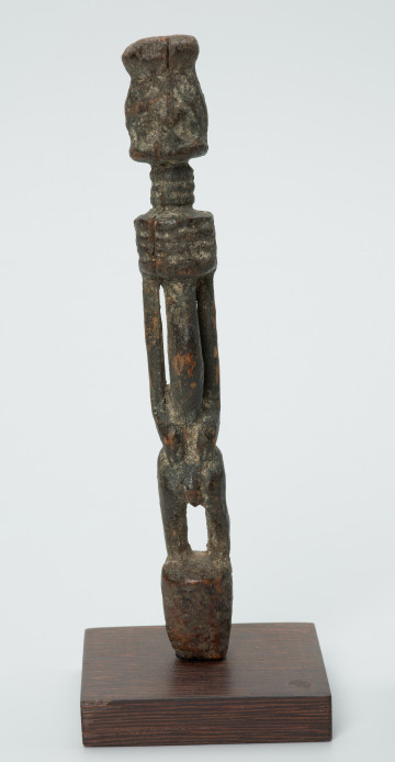 rzeźba - Ujęcie z przodu. Drewniana rzeźba postaci mężczyzny ze zgietymi kolanami. Schematyczne zarysowanie dużej głowy, długiej szyi, na której widoczny jest szeroki naszyjnik. Wąski, długi tułów, długie ręce oparte na kolanach, krótkie nogi i szerokie stopy.