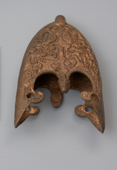 trzewik pochwy miecza typu VI (tzw. grupa gotlandzka) - Ujęcie przodu z góry. Dobrze zachowane dolne okucie pochwy miecza, z niewielkim ubytkiem po jednej stronie i ogólnym wytarciem powierzchni. Na obu bokach widoczne są szwy odlewnicze. Górna część trzewika zaopatrzona jest w trzy występy, dwa boczne ozdobione wypustkami są wyższe od środkowego. Występ środkowy zwieńczony jest stylizowaną główką zwierzęcą. Dolna strona zaopatrzona jest w centralnie umieszczony występ. Obie strony powierzchni trzewika wypełnia skomplikowany ornament, słabo czytelny w wyniku częściowego wytarcia. Przedstawia on drzewo życia z parą stylizowanych ptaków o długich ogonach oraz u podstawy pnia wijące się ósemkowato mocno wydłużone ciała wężów-smoków.