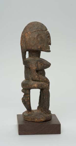 rzeźba - Ujęcie z lewej strony. Figura przedstawiająca postać kobiety siedzącej na stołku. Postać masywna, głowa nieproporcjonalnie duża, ramiona proste, ręce przylegające do tułowia, dłonie spoczywają na kolanach. Nos długi, prosty, w kształcie strzałki, oczy i usta zaznaczone symbolicznie.