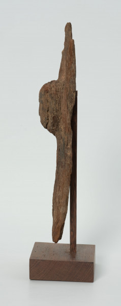 Drewniana figurka na rozwidlonej u góry desce - Ujęcie z lewej strony; na płaskiej, rozwidlonej na szczycie desce wyrzeźbiono okrągłą głowę z delikatnie zaznaczonymi oczami, nosem z małymi ustami. Figura posiada liczne spękania.