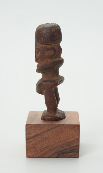 Drewniana figurka postaci ludzkiej z dużą głową i krótkimi nogami. - Ujęcie z lewej strony; drewniana figurka postaci ludzkiej. Głowa zajmuje większość powierzchni rzeźby. Nos wąski, długi, w kształcie strzałki. Brak tułowia. Krótkie nogi.