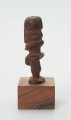 Drewniana figurka postaci ludzkiej z dużą głową i krótkimi nogami. - Ujęcie z lewej strony; drewniana figurka postaci ludzkiej. Głowa zajmuje większość powierzchni rzeźby. Nos wąski, długi, w kształcie strzałki. Brak tułowia. Krótkie nogi.