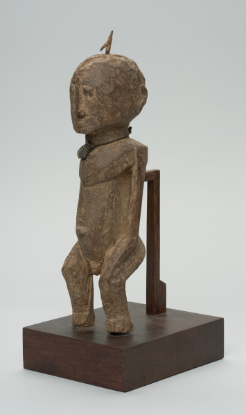 rzeźba - Ujęcie z przodu z lewej strony. Figura przedstawiająca mężczyznę, o czym świadczy lekko zaznaczony penis. Prawa ręka krótsza od lewej, uniesiona do góry. Ramiona proste, pośladki i uda zaokrąglone. Na głowie wbity kawałek żelaza. Związana z kultem przodków.
