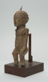 rzeźba - Ujęcie z przodu z lewej strony. Figura przedstawiająca mężczyznę, o czym świadczy lekko zaznaczony penis. Prawa ręka krótsza od lewej, uniesiona do góry. Ramiona proste, pośladki i uda zaokrąglone. Na głowie wbity kawałek żelaza. Związana z kultem przodków.